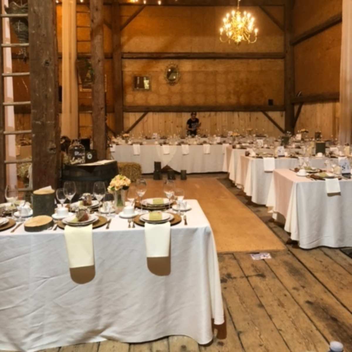 A banquet hall features long tables set with white tablecloths, glassware, and dining utensils, under warm lighting from chandeliers and string lights.