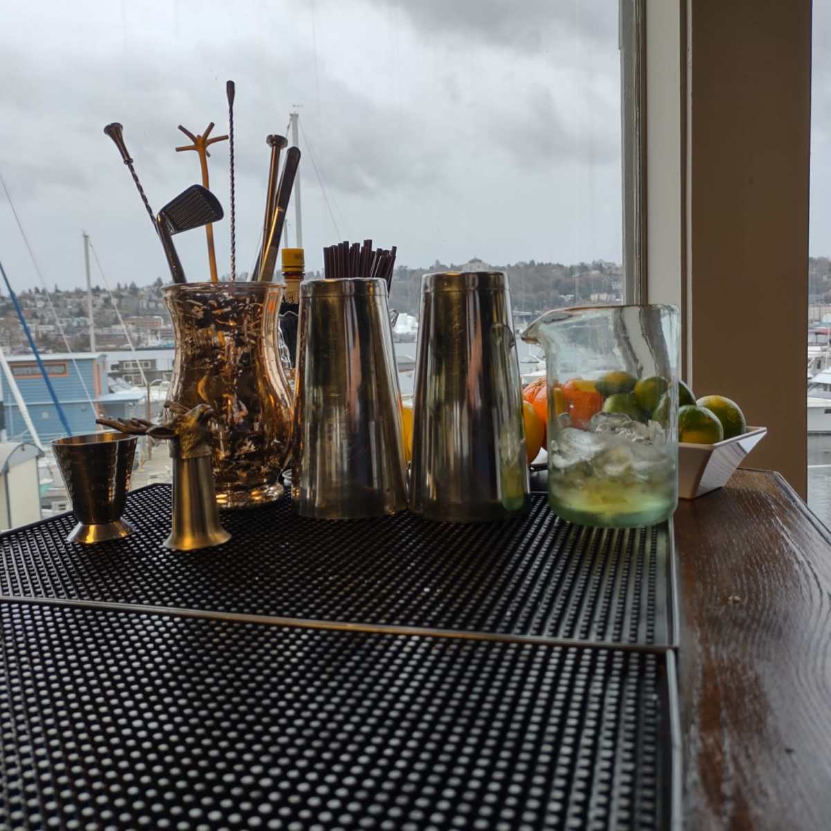 A bar counter displays various cocktail tools and a pitcher next to a glass of drink, with boats visible outside the window.