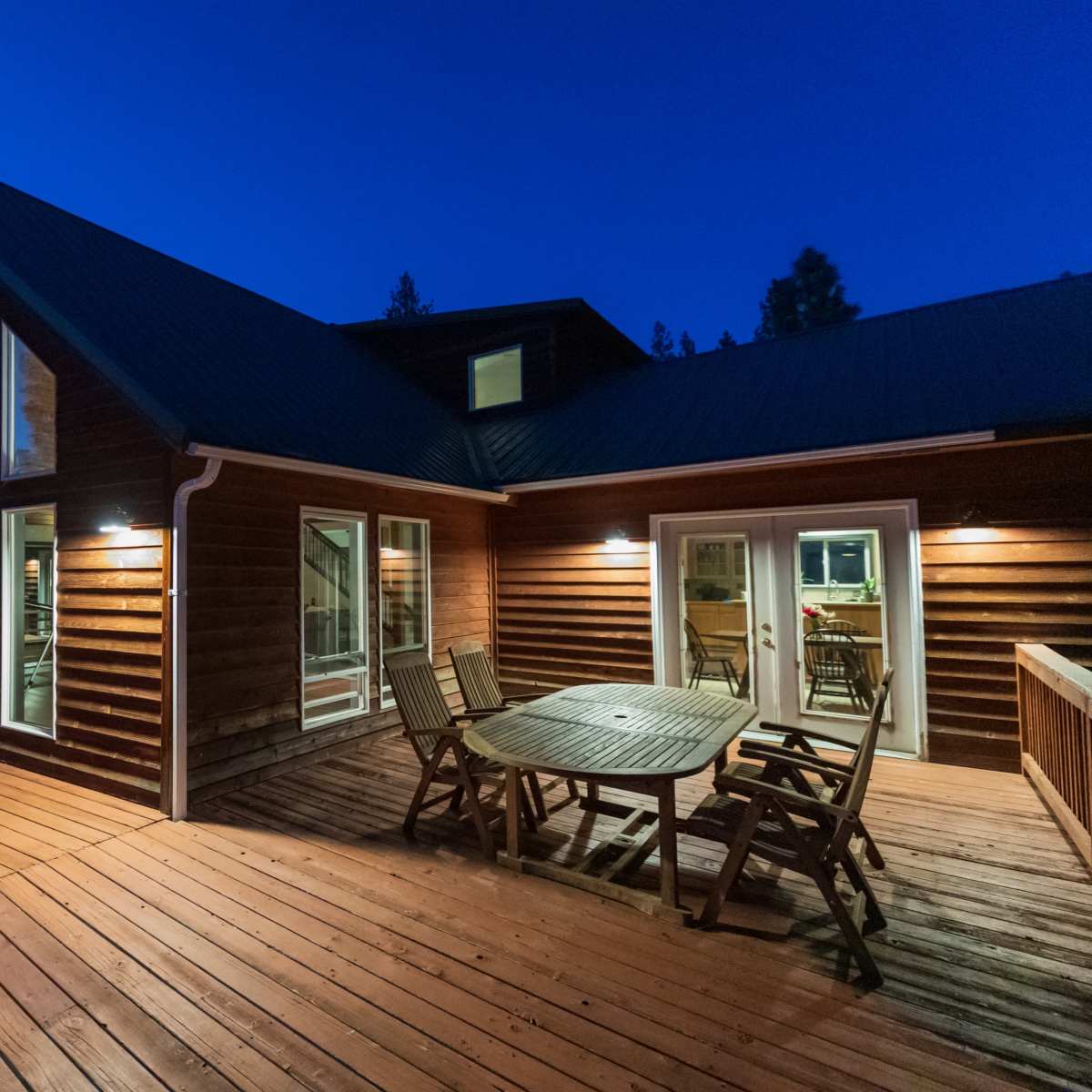 A wooden deck surrounds a modern house with large windows, situated in a wooded area at dusk.