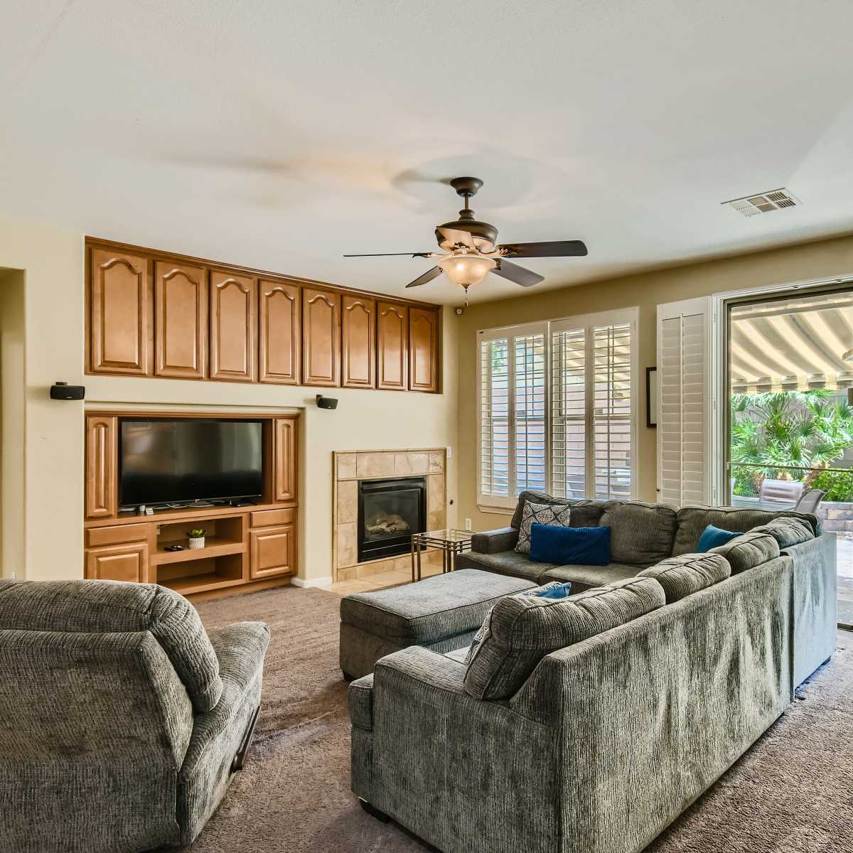 The living room features a ceiling fan, a wall-mounted television, a fireplace, and large windows leading to an outdoor patio.