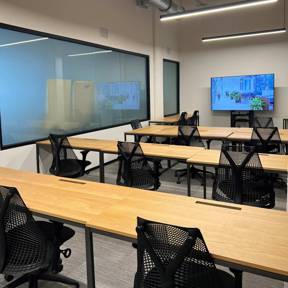 A modern conference room featuring several wooden tables arranged for a meeting, surrounded by black mesh chairs and a large screen on the wall.
