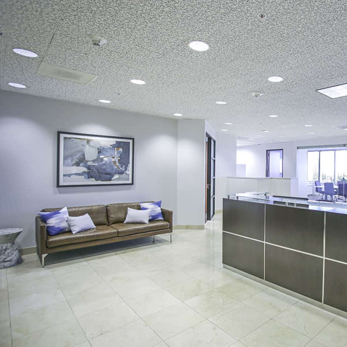 A modern office reception area with a brown sofa, a reception desk, and large windows allowing natural light in.