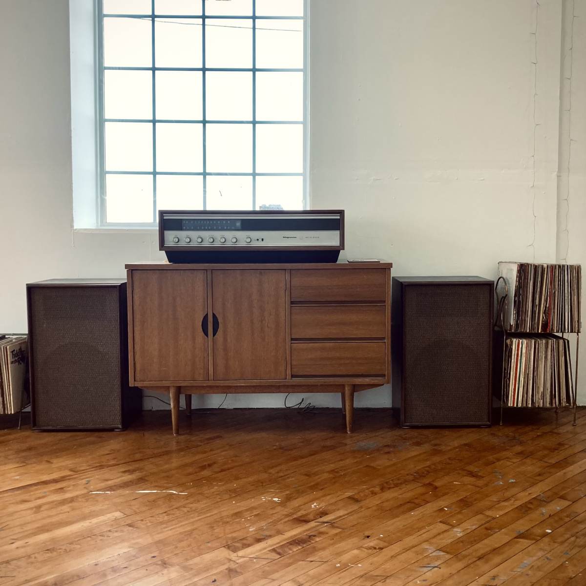 A vintage stereo system is positioned on a wooden cabinet, flanked by two speaker units and stacks of vinyl records against a windowed wall.