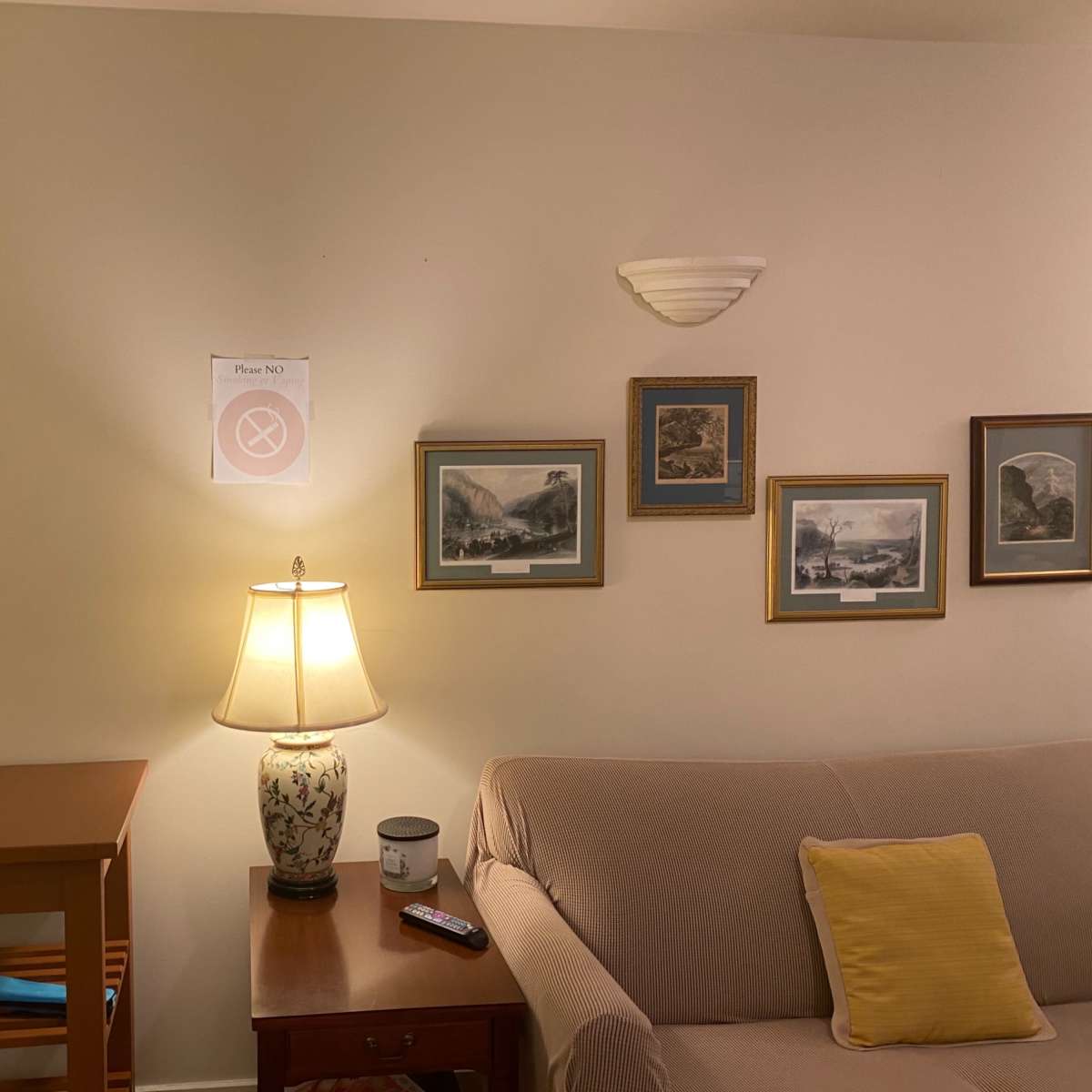 A beige sofa sits beside a wooden side table with a lamp, while framed pictures line the wall, and a sign indicating "Please Do Not" hangs above.