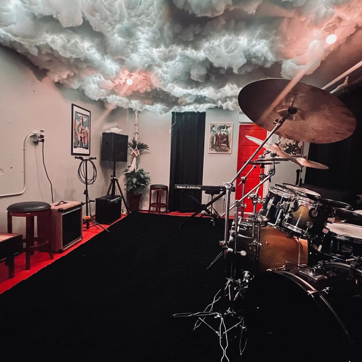 A music rehearsal space with a drum set, keyboard, and sound equipment, featuring a cloud-like ceiling and black carpet.