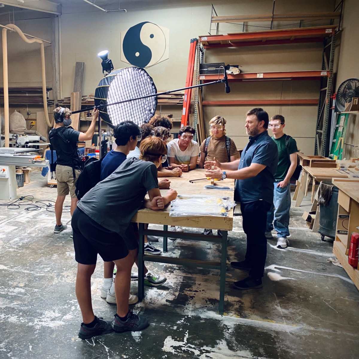 A group of people gathers around a workbench in a workshop while a camera crew films the scene.