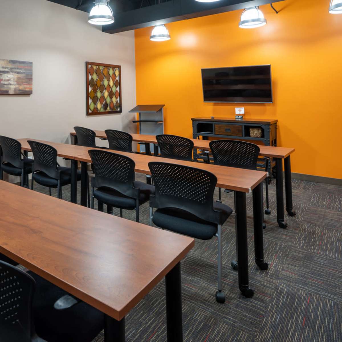 A classroom with long wooden tables and black chairs is set up for a training or meeting, featuring an orange accent wall and a TV on a media console.