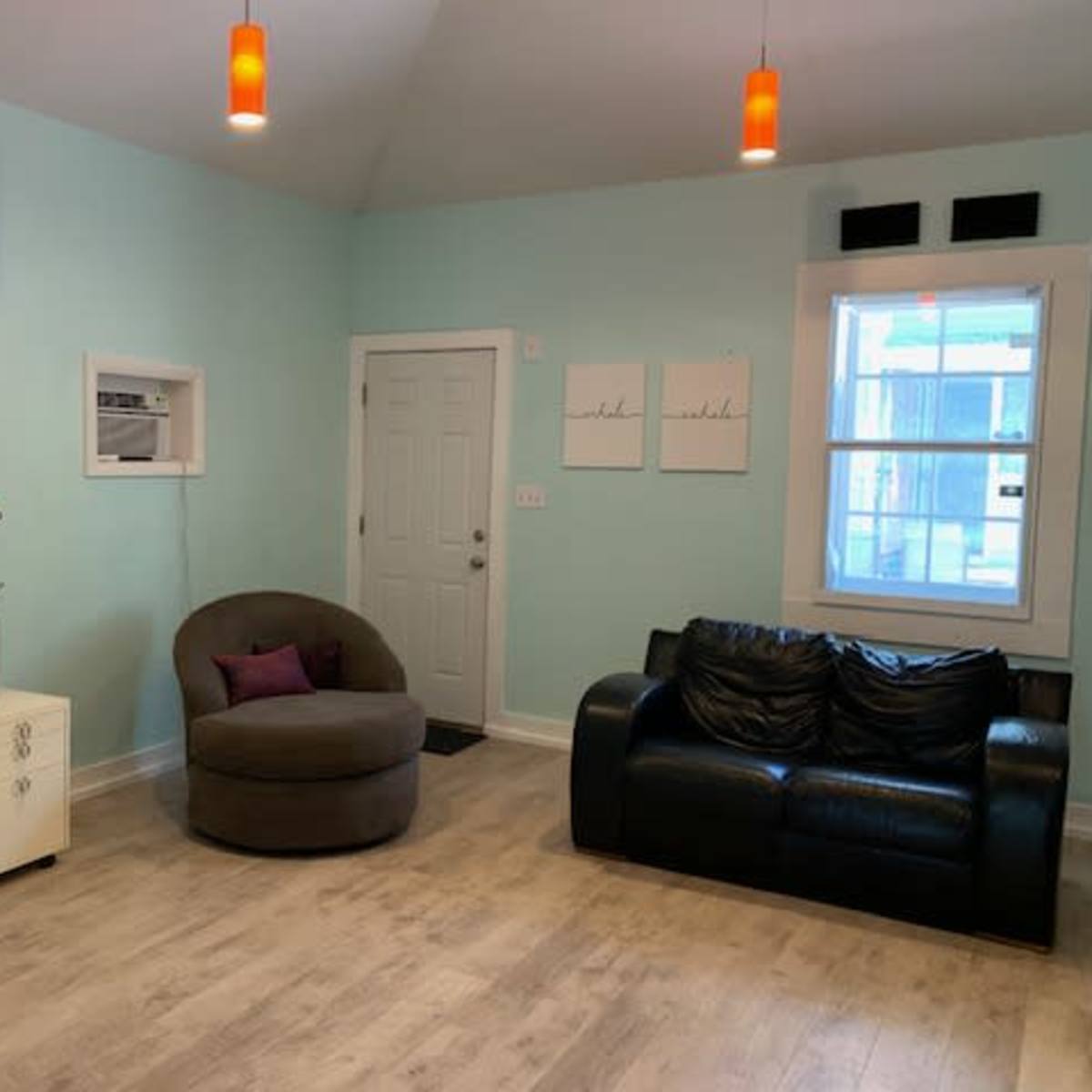 A light blue room with a brown armchair, a black leather sofa, a white wall-mounted air conditioning unit, and two framed artworks on the wall.