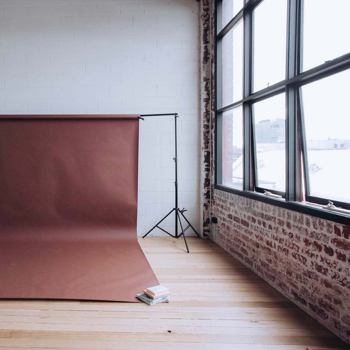 A photography backdrop stands on a frame in a spacious room with large windows, and two stacks of books are placed on the floor at the base.
