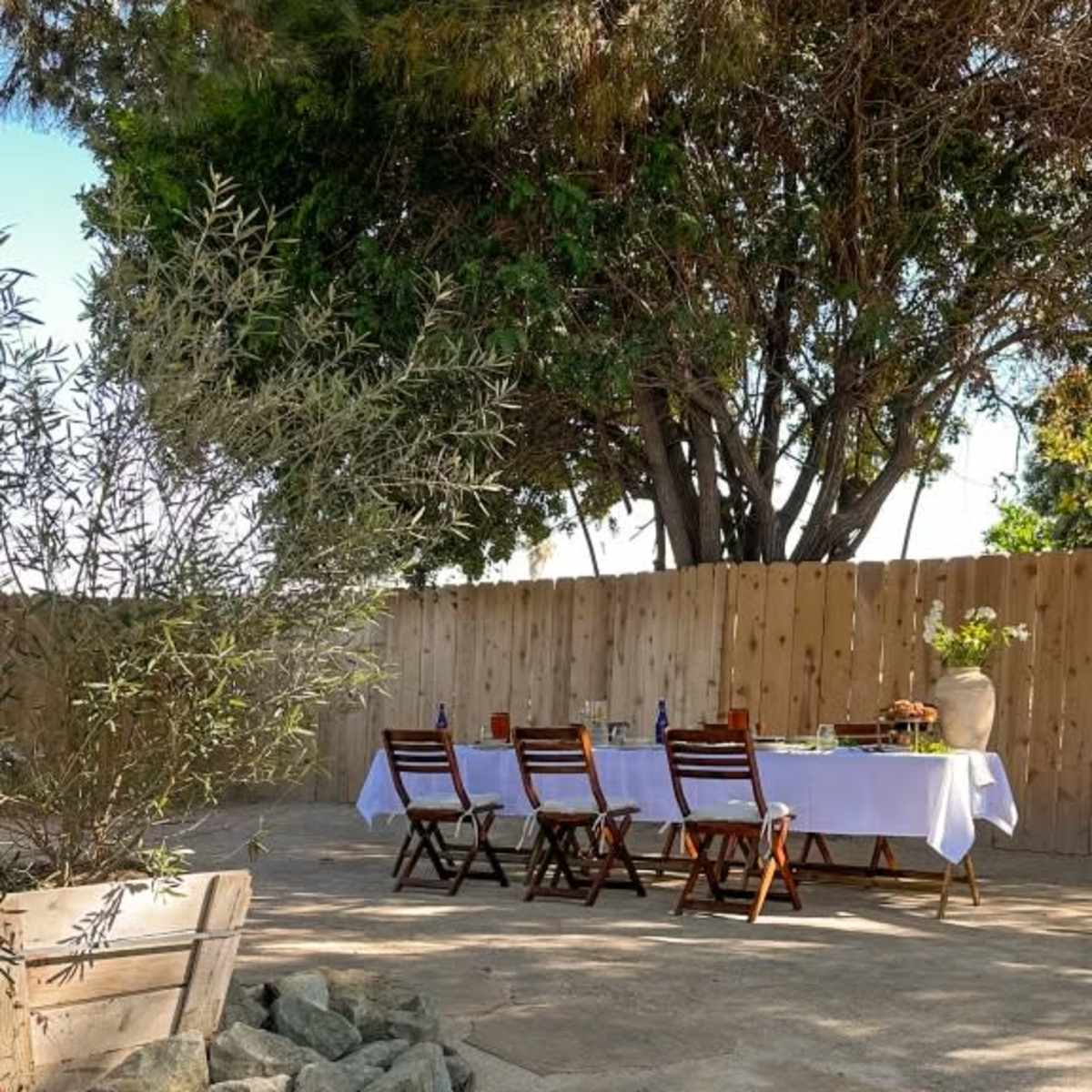 A long dining table is set outdoors with chairs, surrounded by trees and a wooden fence.