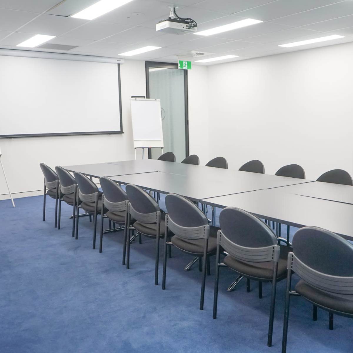 A conference room features a long table surrounded by black chairs, with a projector screen and whiteboards in the background.