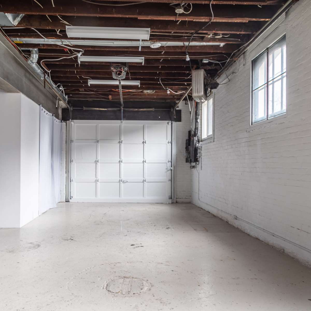 An empty garage with a white wall on one side, a garage door, and exposed ceiling beams.