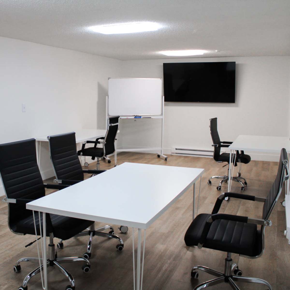 A minimalist meeting room with four black office chairs and two white tables, featuring a wall-mounted television and a whiteboard.