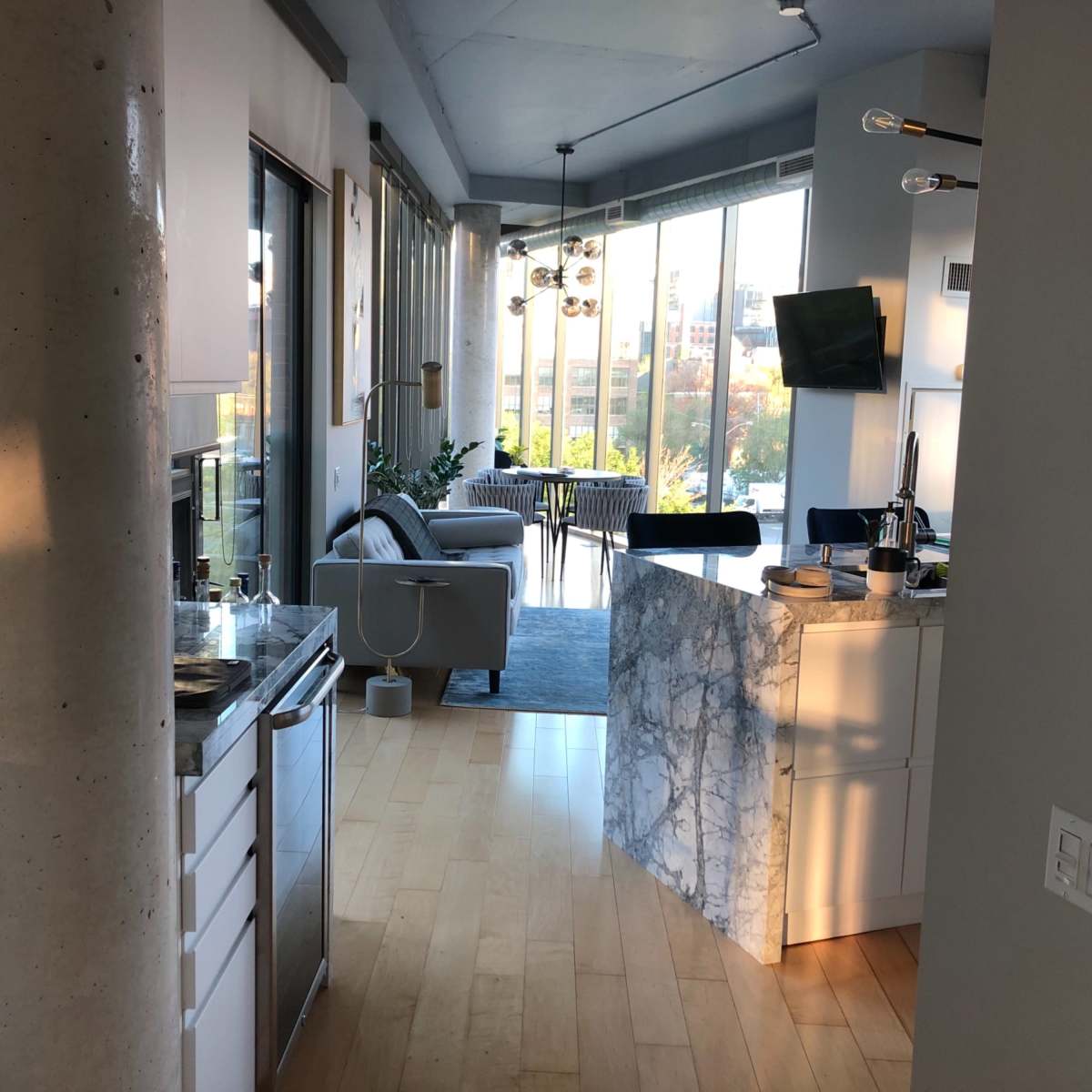 A modern open-concept kitchen and living area with large windows allowing natural light, featuring a marble kitchen island and contemporary furnishings.