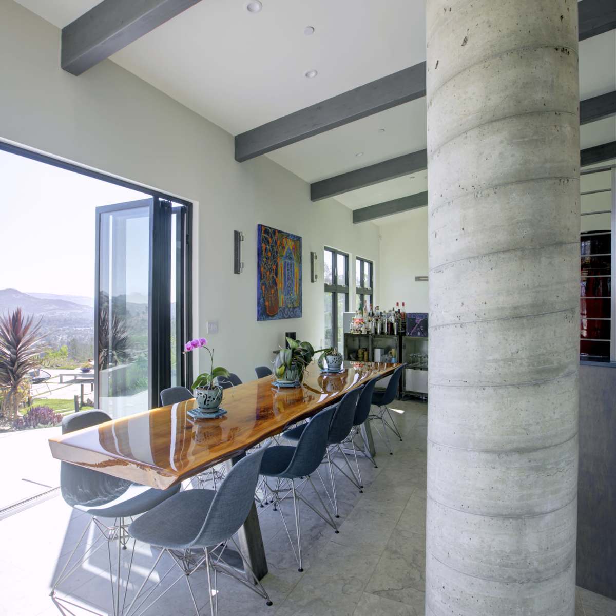 A long wooden dining table with gray chairs is positioned near a large opening that leads to an outdoor view, with a concrete pillar supporting the ceiling.