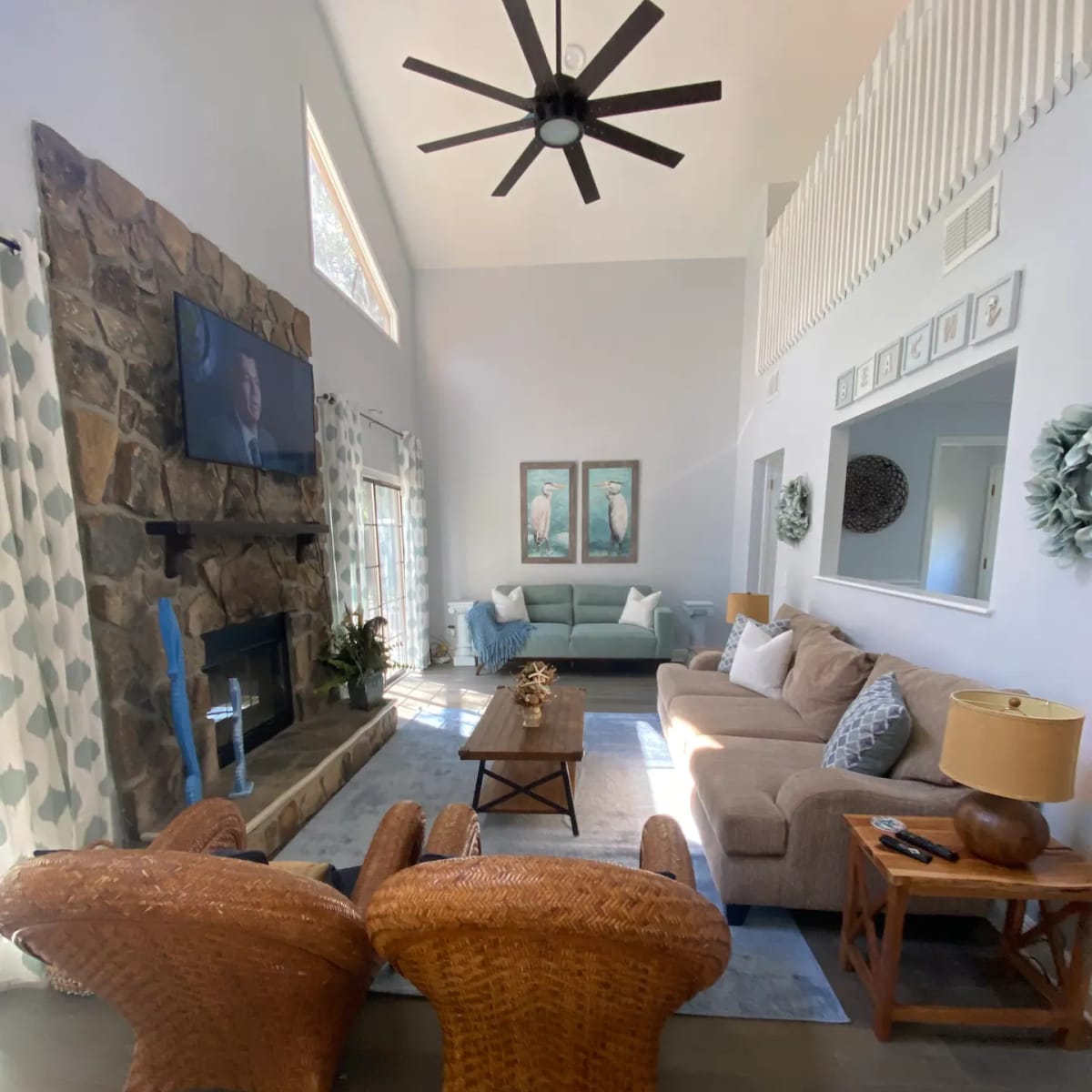 A spacious living room featuring a stone fireplace, a large sofa, two woven armchairs, and a television mounted on the wall.