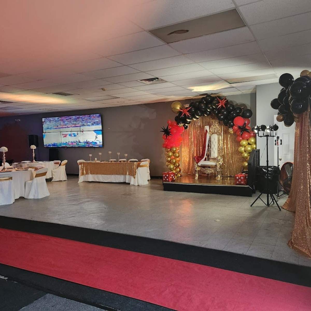 An event space set up for a celebration, featuring a stage with gold and black balloons, a decorative backdrop, and tables arranged for guests.