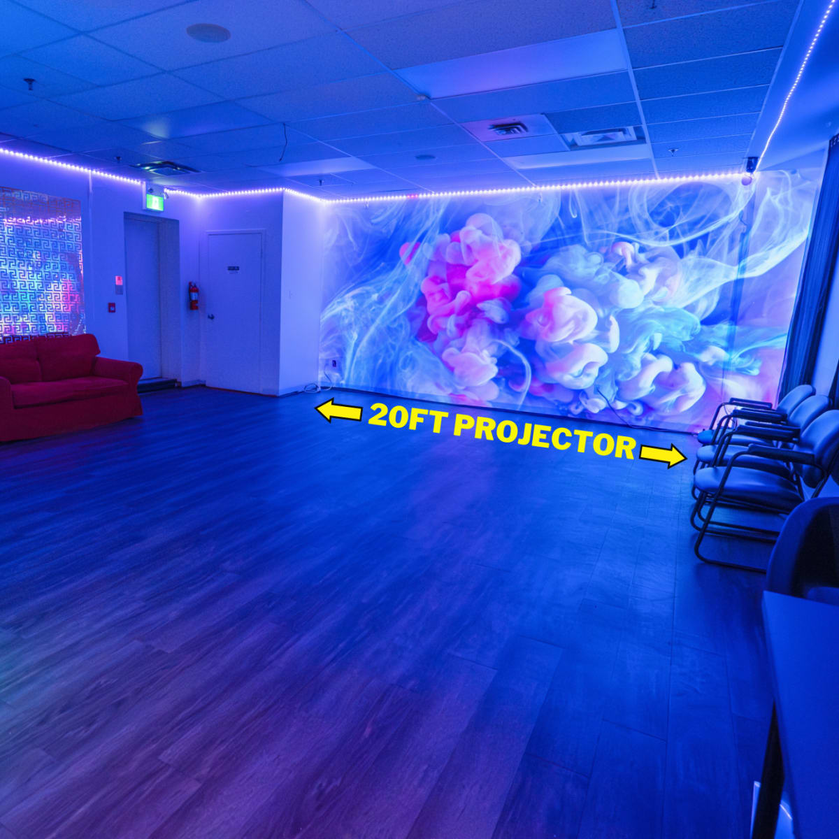A spacious, brightly lit room features a red sofa, various chairs, and a large wall with projected colorful visuals.