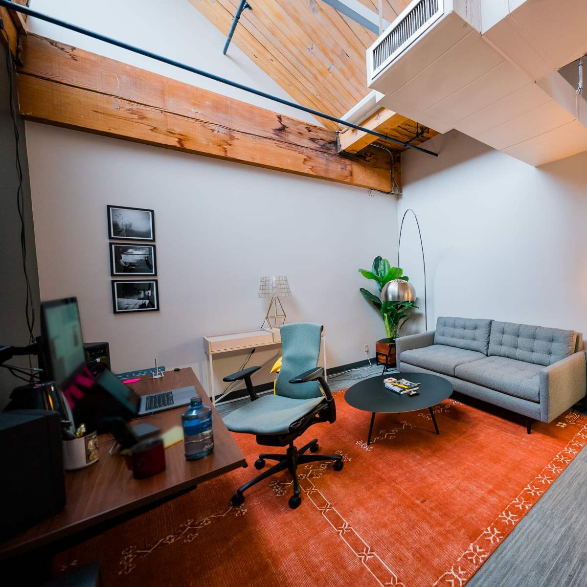 A modern office space features a gray sofa, a round black coffee table, a desk with an ergonomic chair, and various decorative elements including a plant and a warm light source.