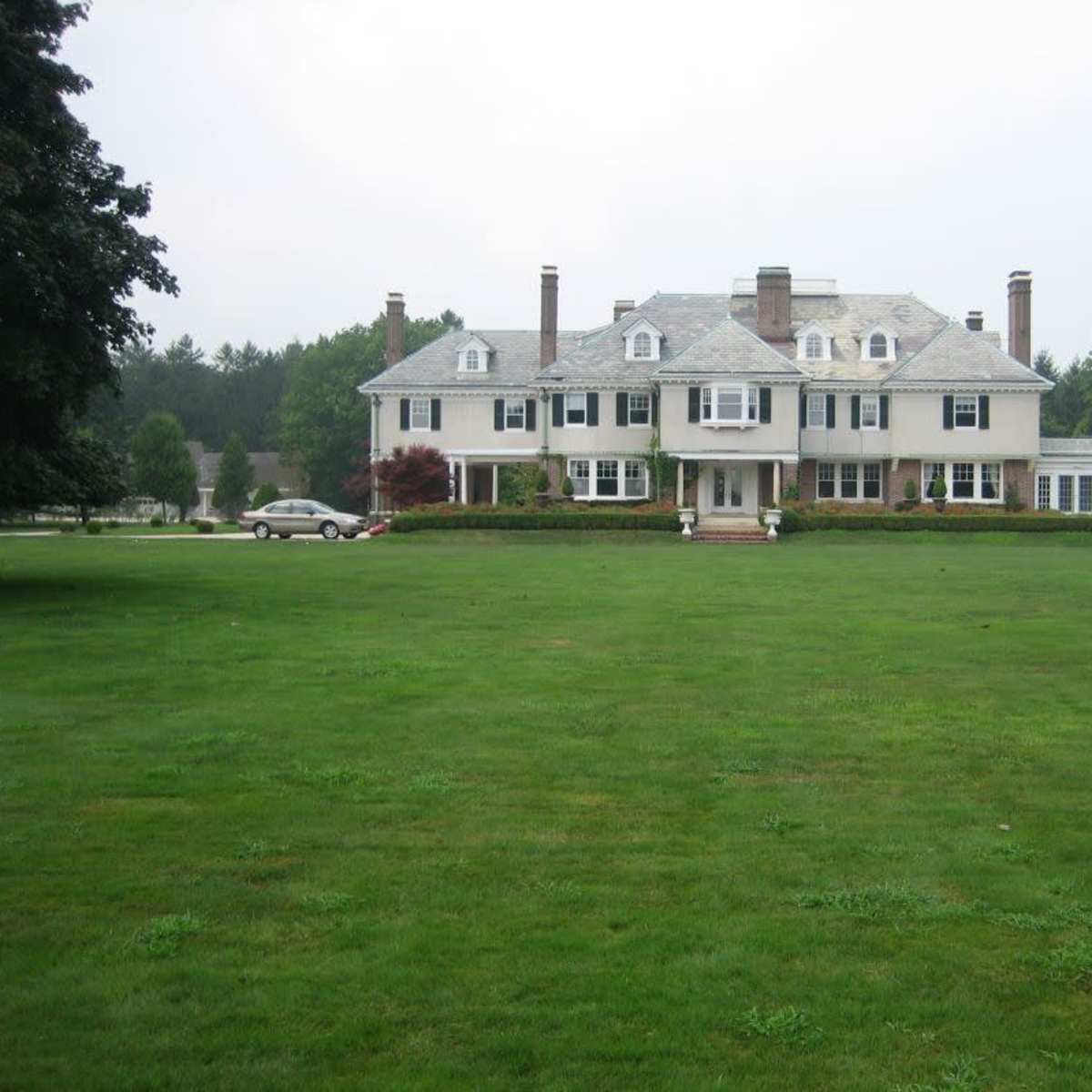 A large, two-story house with a gray roof and white exterior sits on a sprawling green lawn.