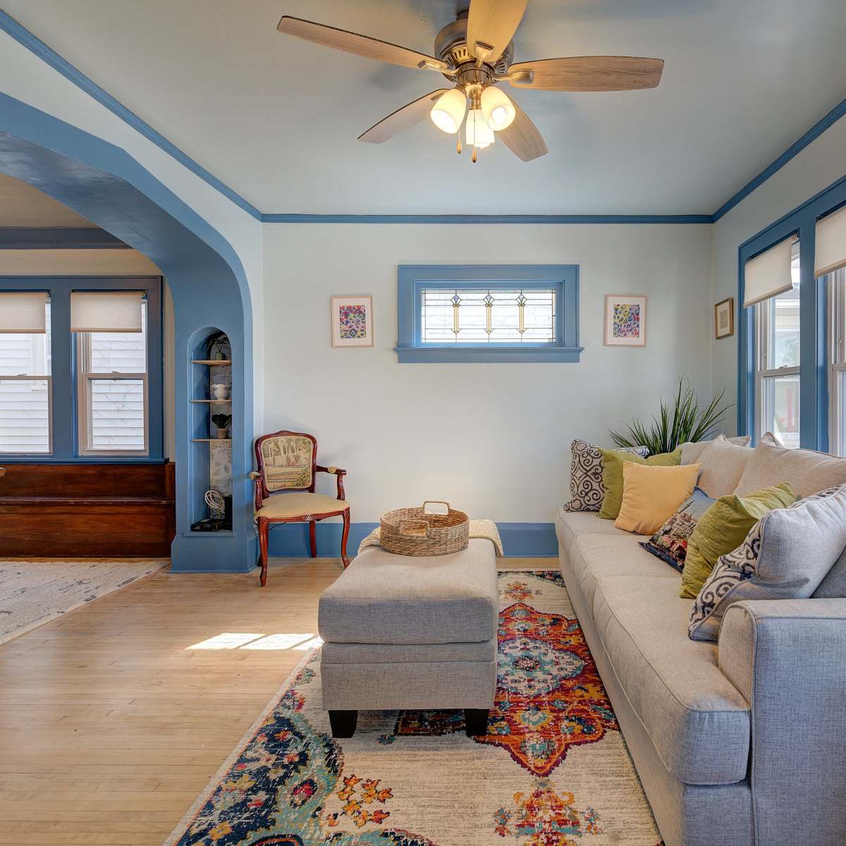 A living room with a light blue color scheme, featuring a beige sofa, an ottoman, decorative pillows, and large windows that allow natural light to illuminate the space.
