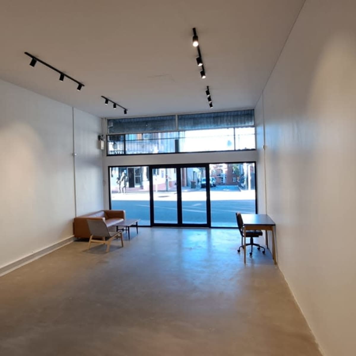 An empty interior space with a concrete floor, two pieces of furniture, and large windows facing a street.