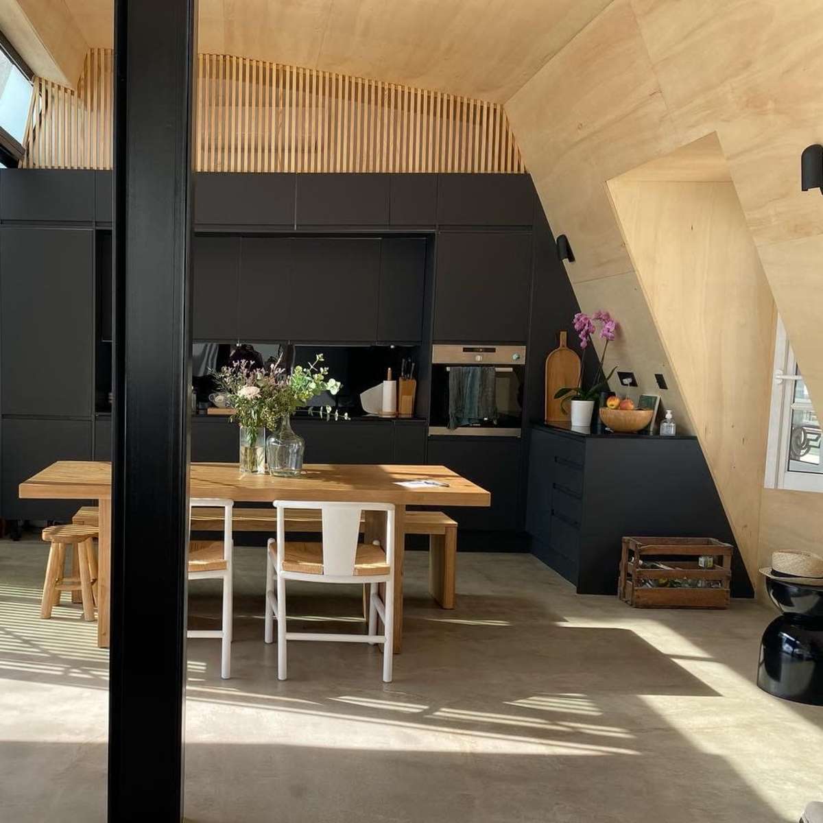 A modern kitchen with an angled wooden ceiling, a large dining table, and sleek black cabinets, illuminated by natural light from large windows.