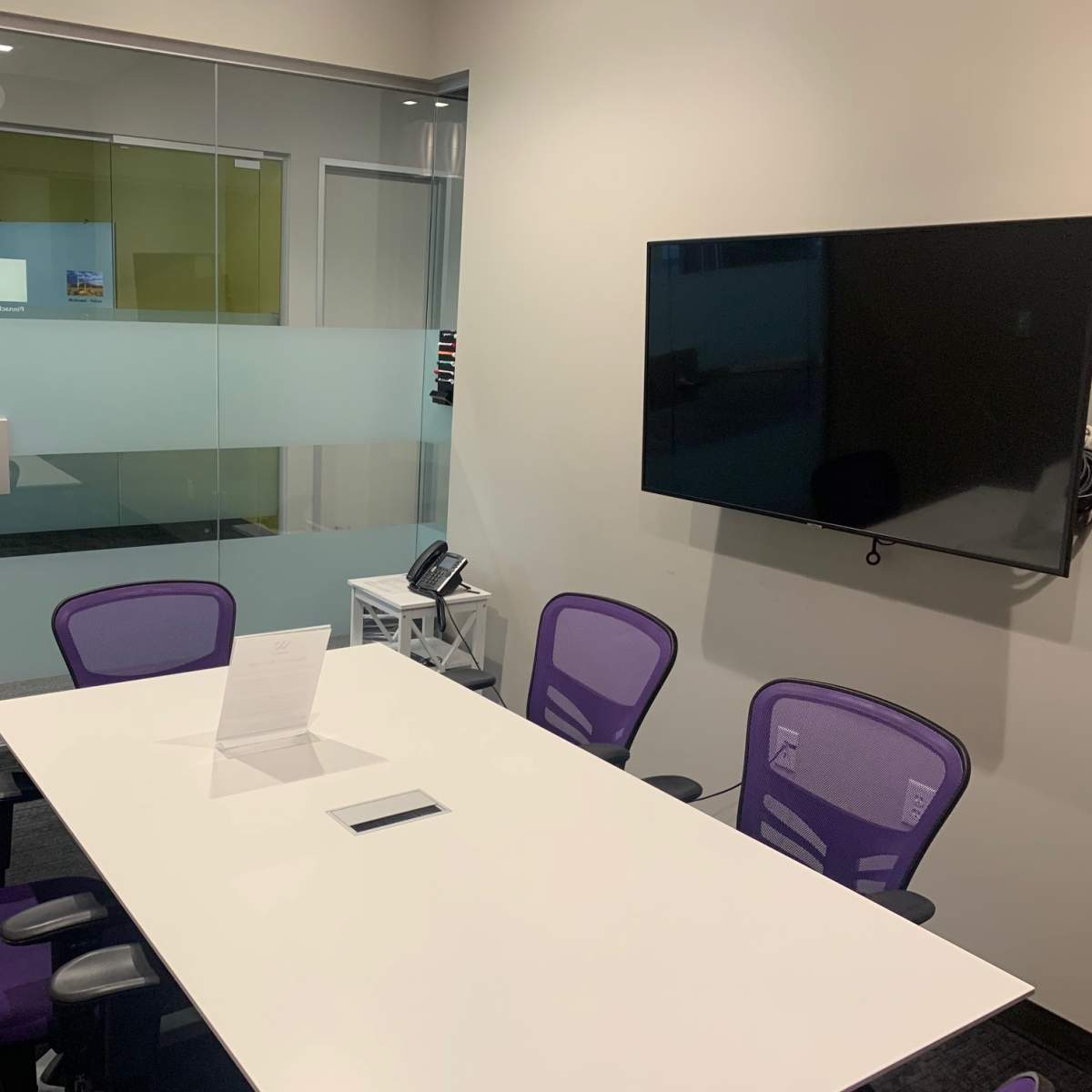 A meeting room features a white table surrounded by purple chairs and a mounted television on the wall.