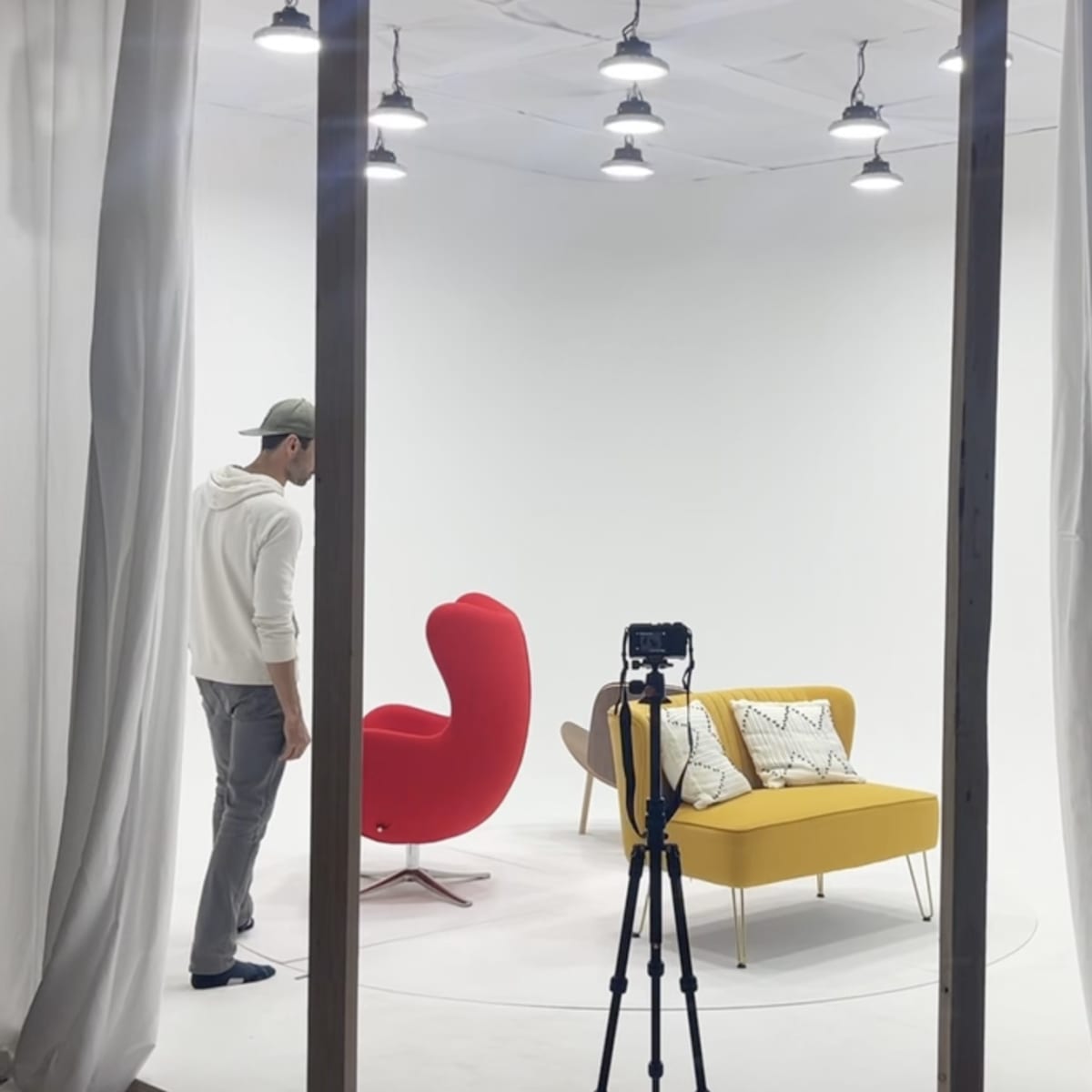 A person stands in a minimalistic studio with two brightly colored chairs and a camera on a tripod.