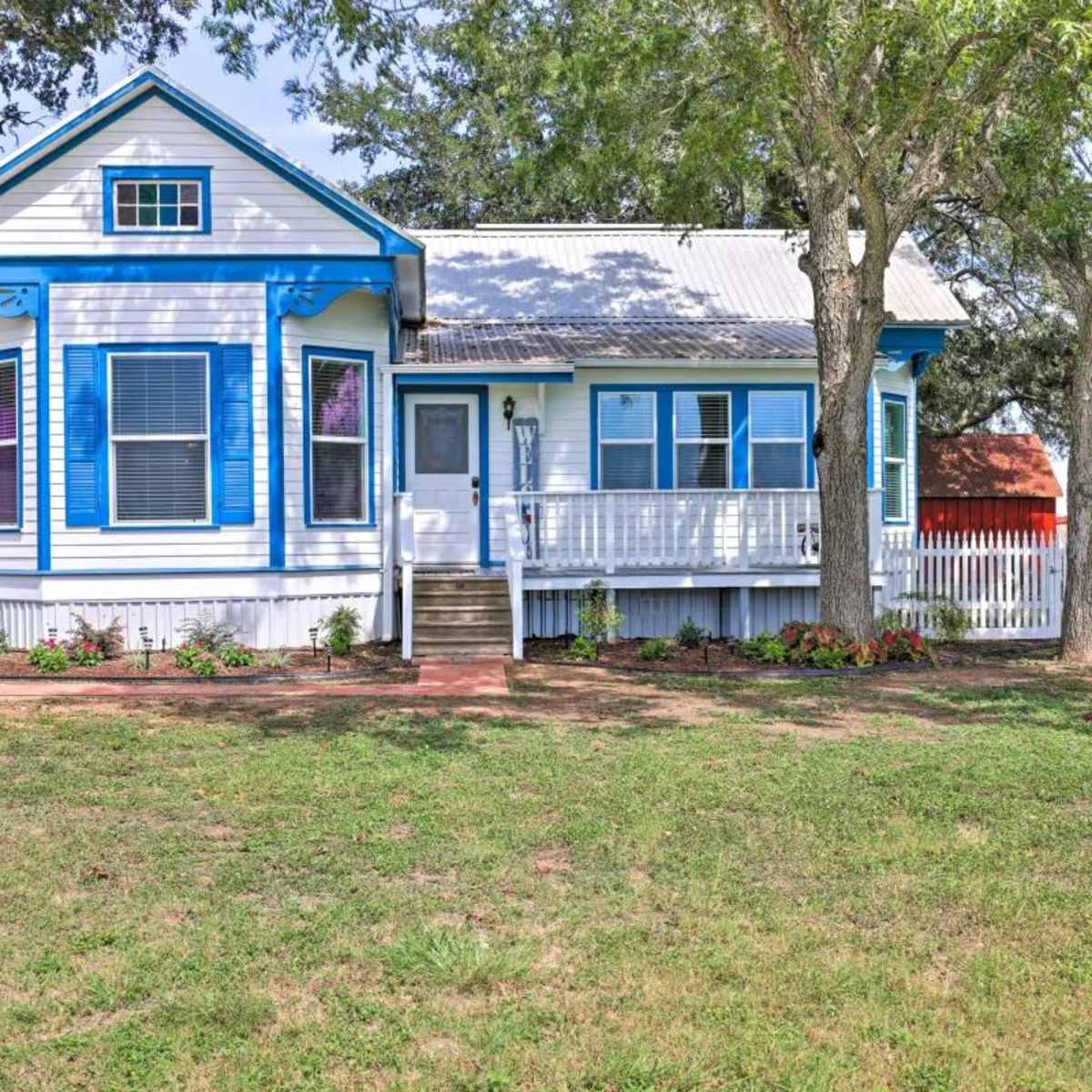 A white house with blue trim and a front porch is surrounded by green grass and trees, with a white picket fence in the background.