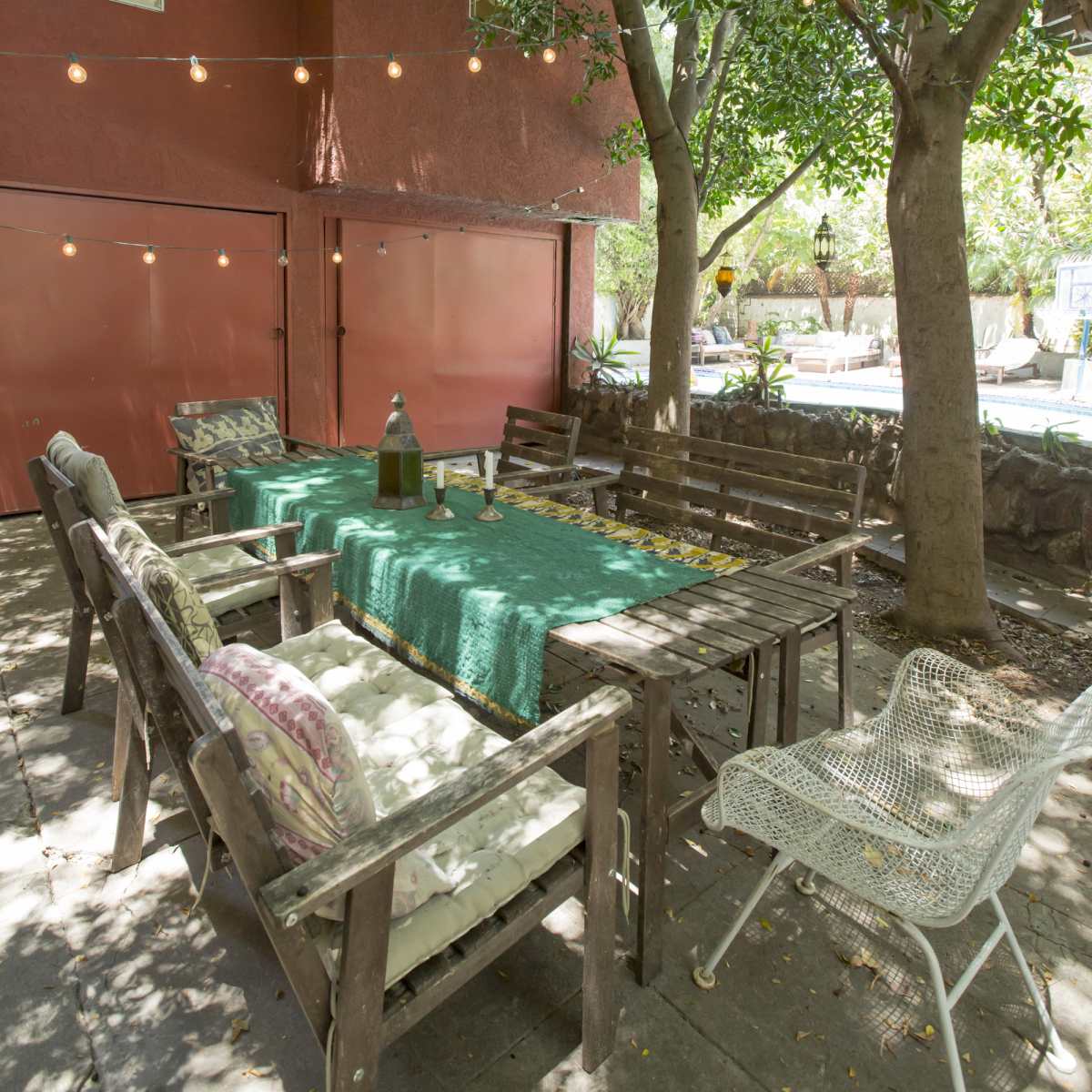 A wooden outdoor dining table is set under trees with string lights, surrounded by chairs in a shaded area near a red building.