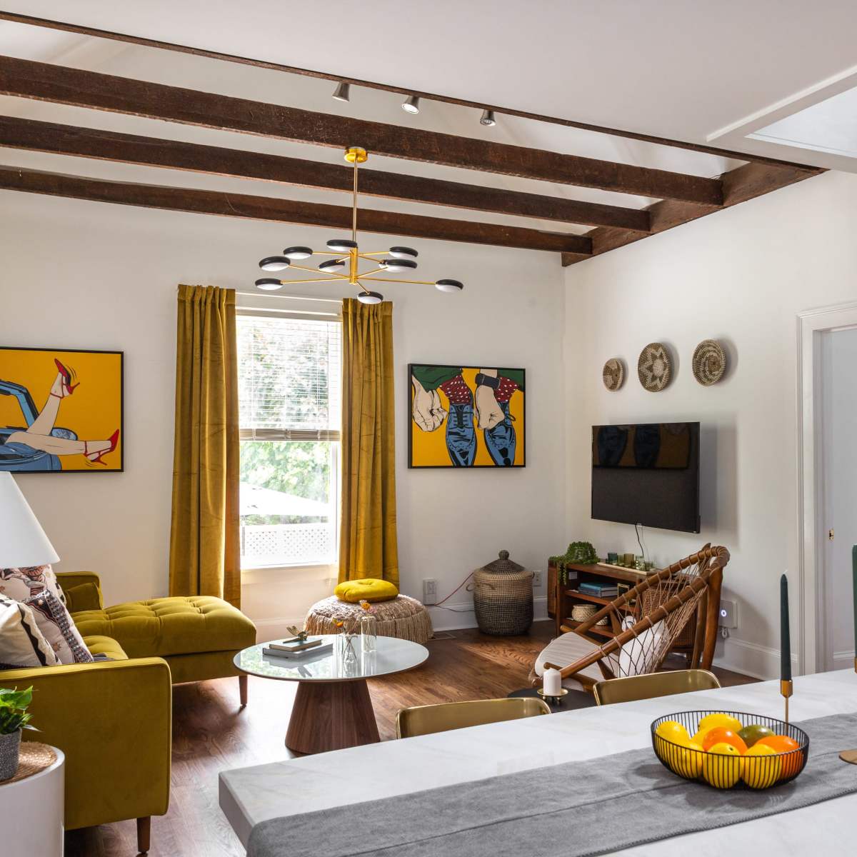 A bright and modern living room features wooden beams, yellow curtains, and contemporary artwork, with a glass coffee table and a spiral staircase leading to an upper level.