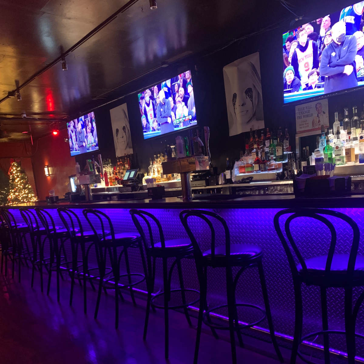 A dimly lit bar with several monitors displaying a basketball game, illuminated by blue LED lights along the counter.