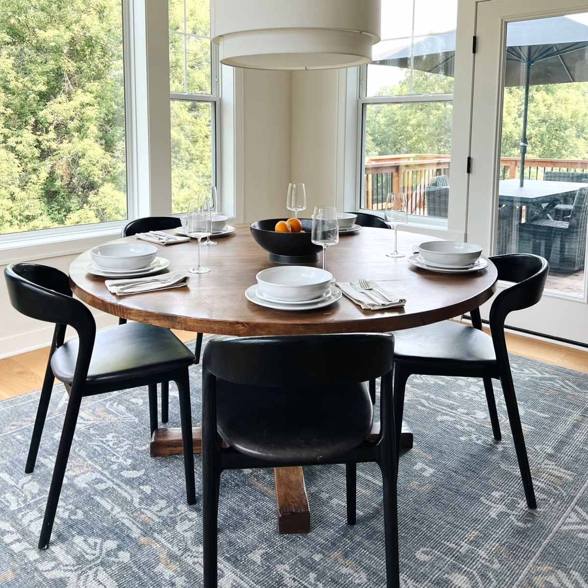 A round wooden dining table is set with tableware and a bowl of fruit, surrounded by black chairs, in a bright room with large windows overlooking greenery.
