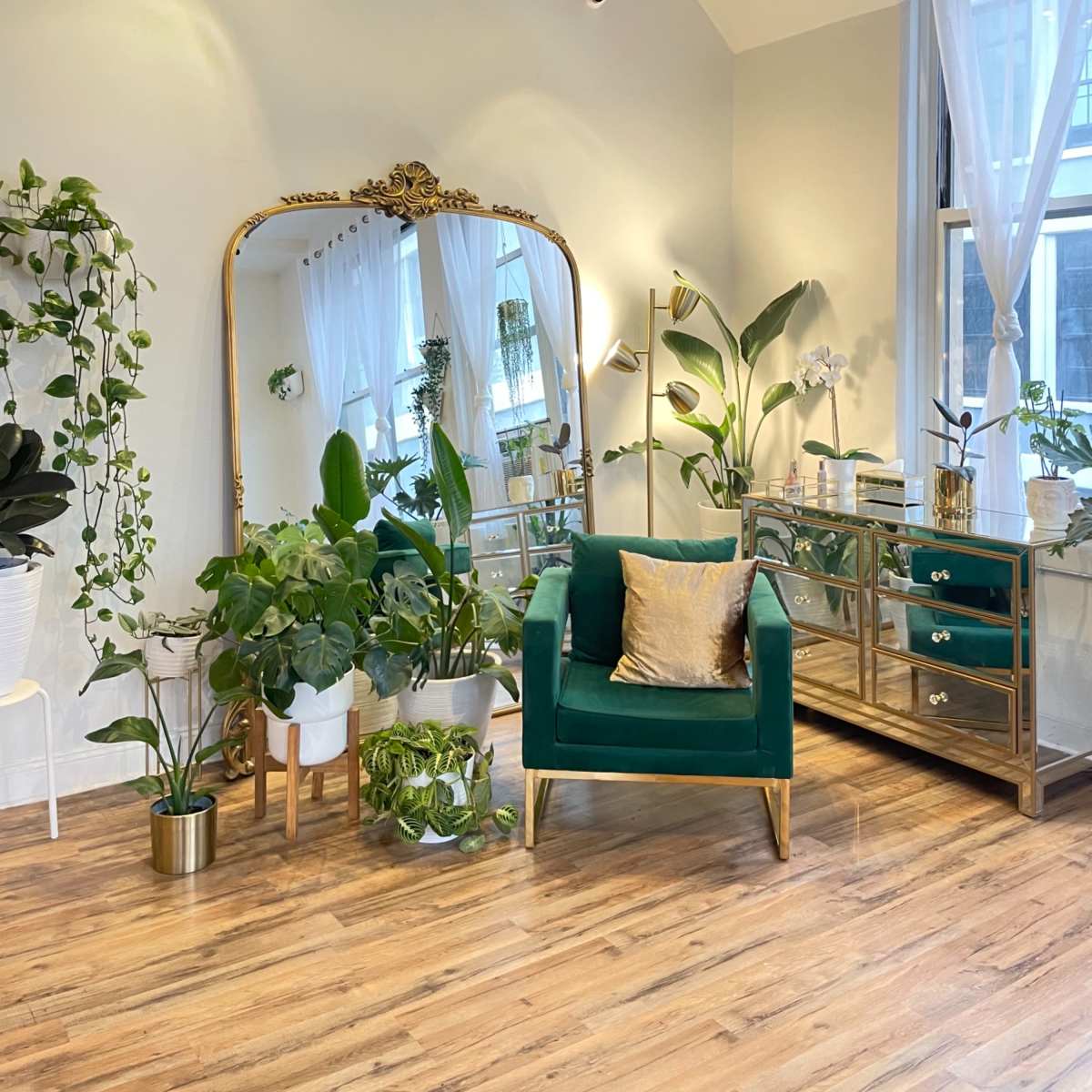 A cozy room featuring a green velvet armchair, a large gilded mirror, and a variety of indoor plants arranged around a gold and glass console table.