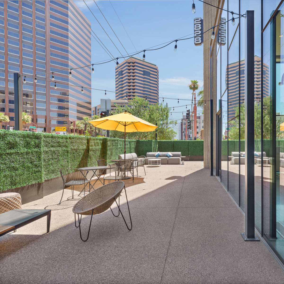 A patio area features outdoor furniture and yellow umbrellas, surrounded by green hedges and buildings in the background.