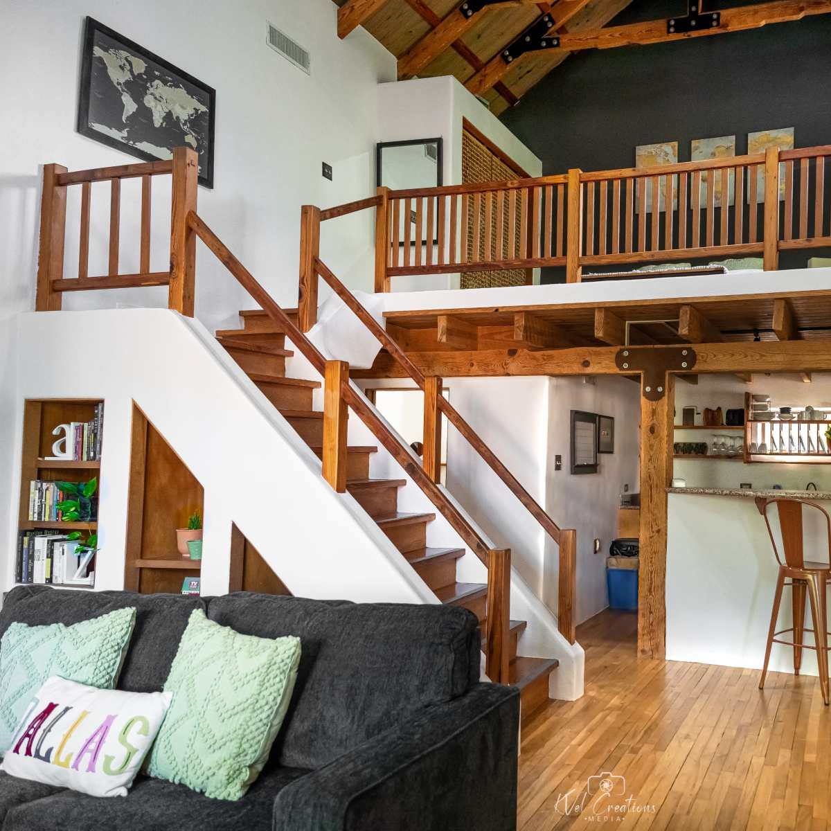 A spacious interior of a house with a loft, featuring a staircase, wooden beams, a seating area with a gray couch, and a bar with two stools in the background.