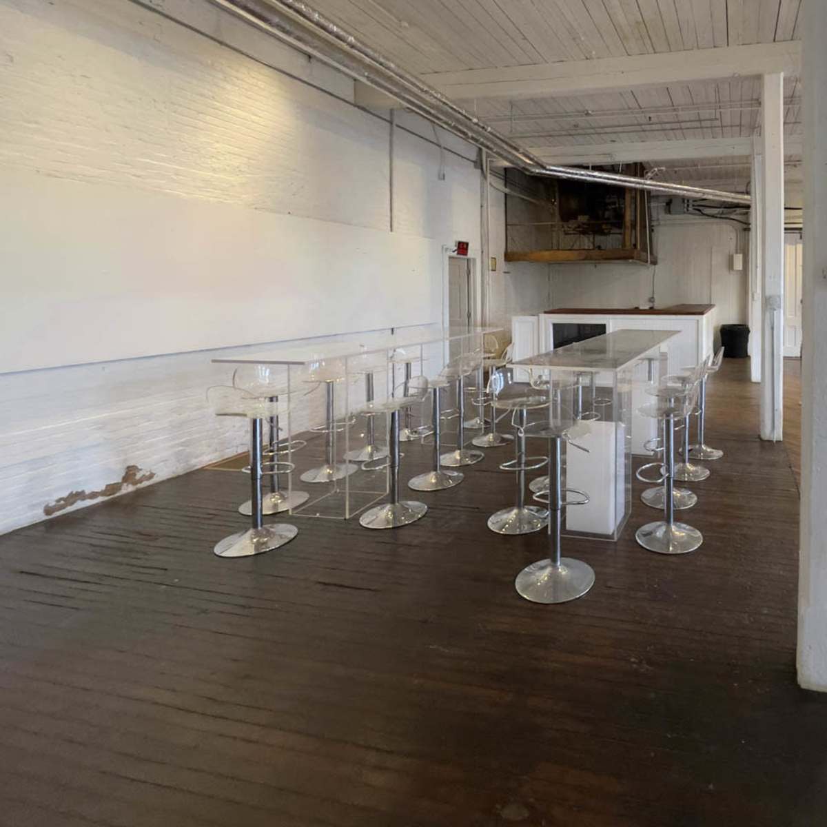 A spacious, minimalist interior with clear acrylic stools arranged around white tables in an industrial-style setting.