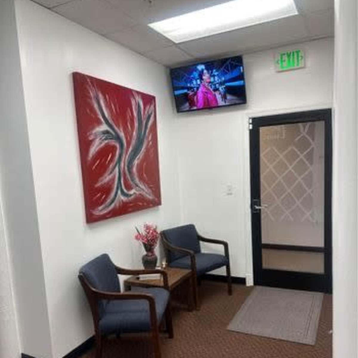 A small waiting area with two chairs, a side table, a piece of abstract art on the wall, and a TV mounted above displaying a program.