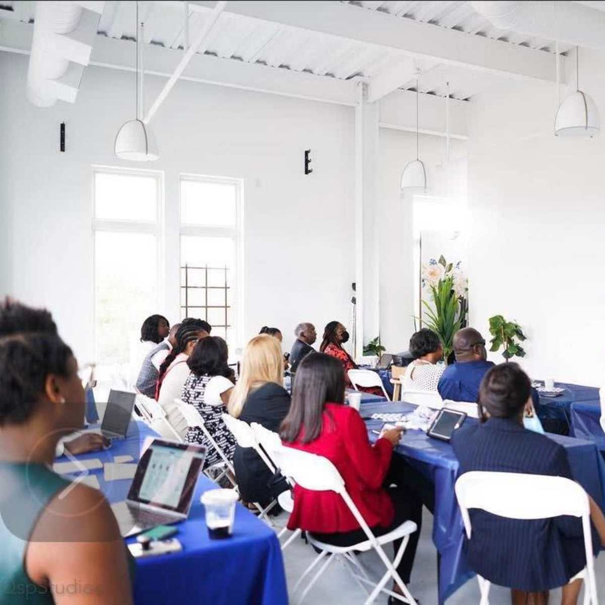 A group of people sits at tables in a bright room, facing a speaker who is presenting at the front.