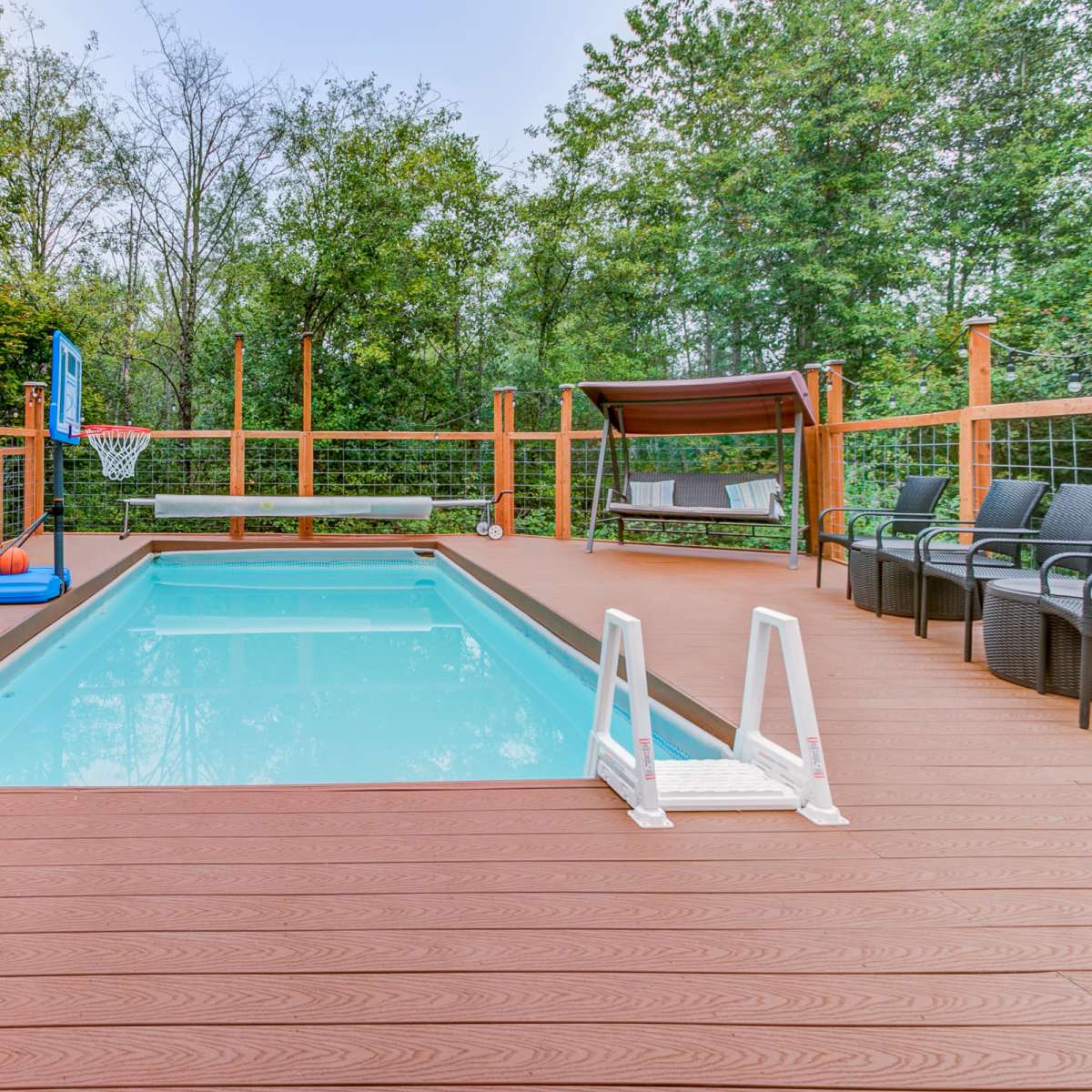 A swimming pool surrounded by a wooden deck, with a basketball hoop, seating area, and a shaded canopy nearby.