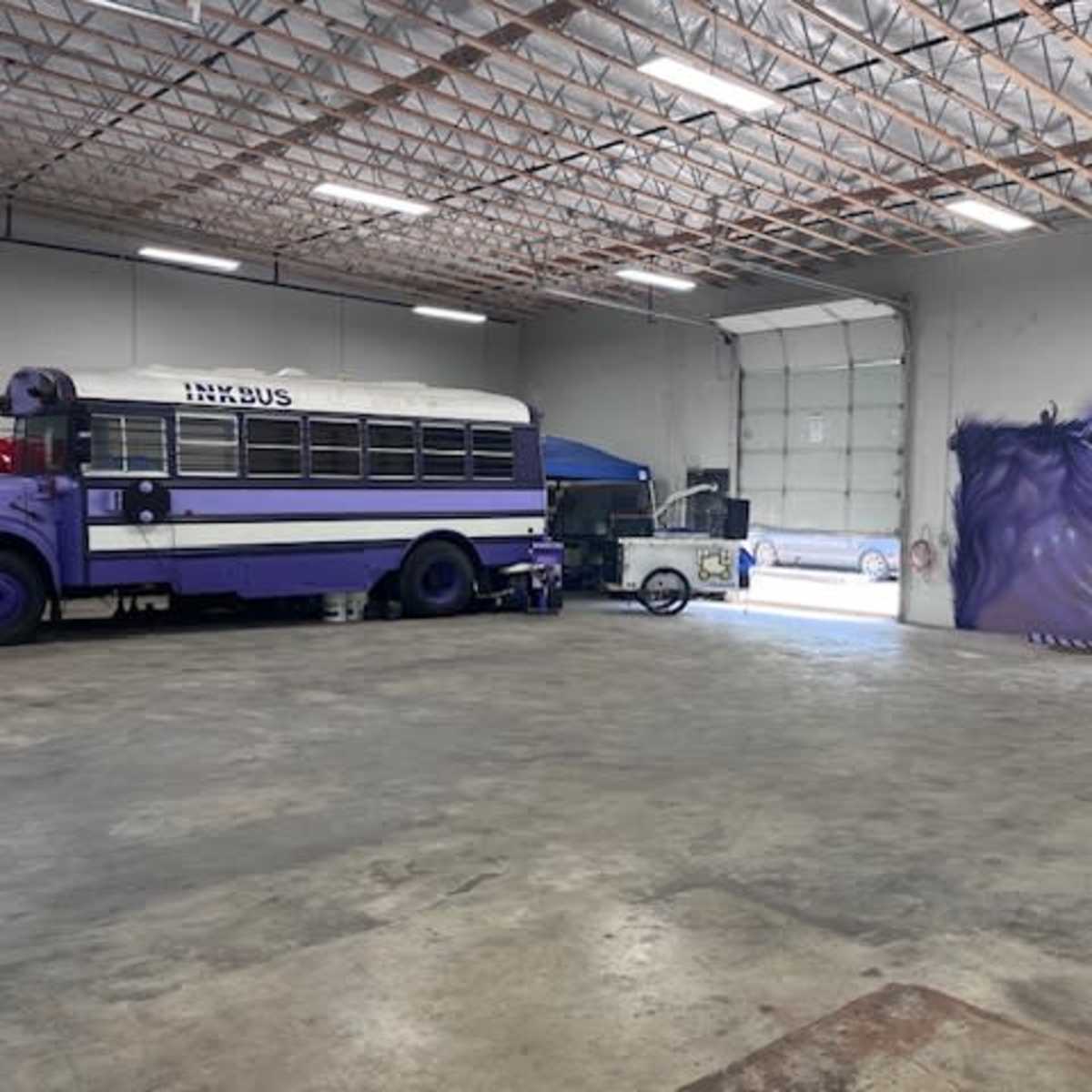 A purple school bus is parked inside a spacious industrial garage, with a mural of an eye on the gray wall.