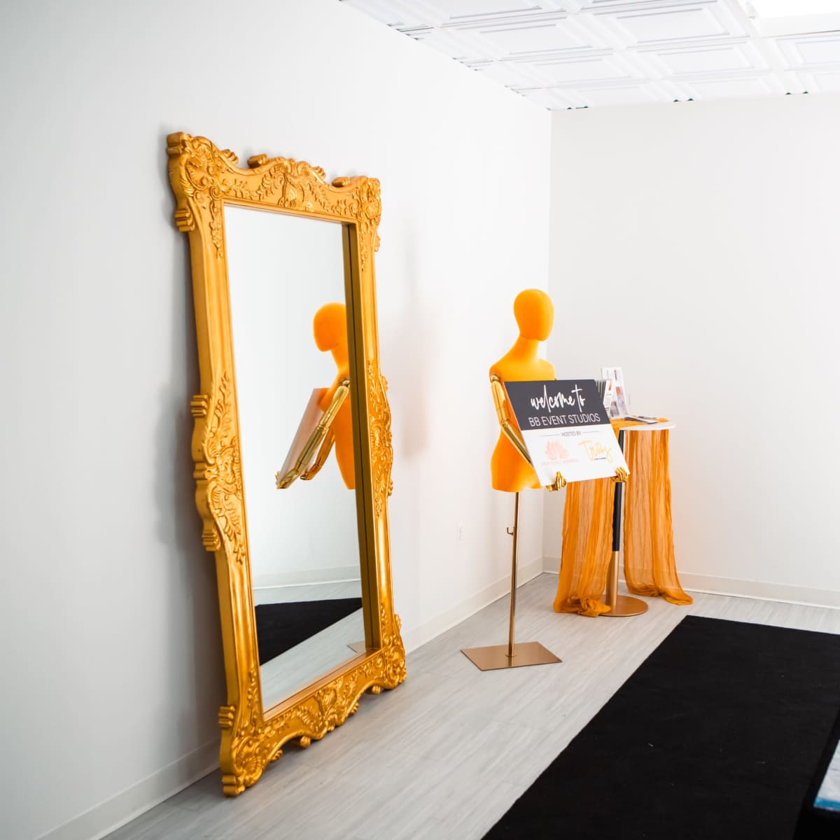 A large ornate gold mirror, a bright orange mannequin, a small display table with a sign, and a fire extinguisher on a white wall background.