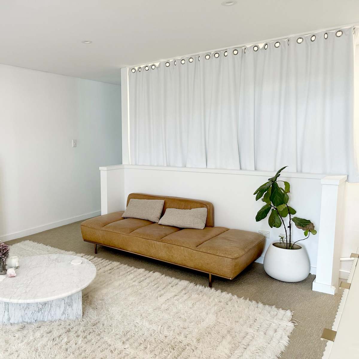 The living room features a brown leather sofa, a round marble coffee table, and a potted plant beside a white curtain, set against a neutral backdrop.