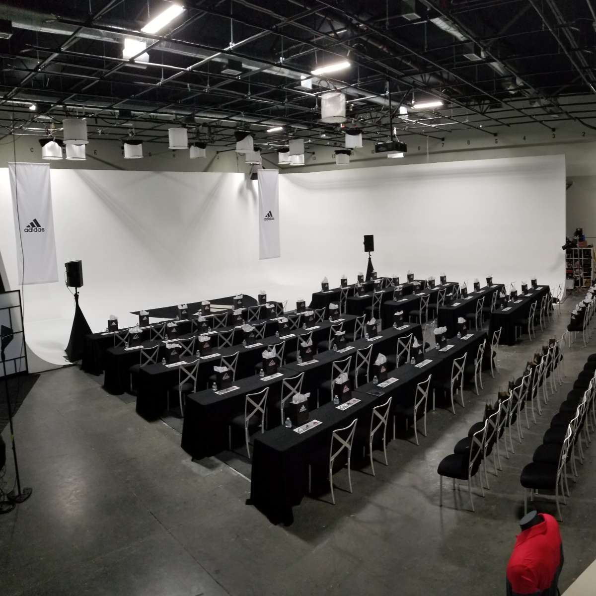 A large empty event space is set up with rows of tables and chairs, flanked by white walls and an Adidas banner.