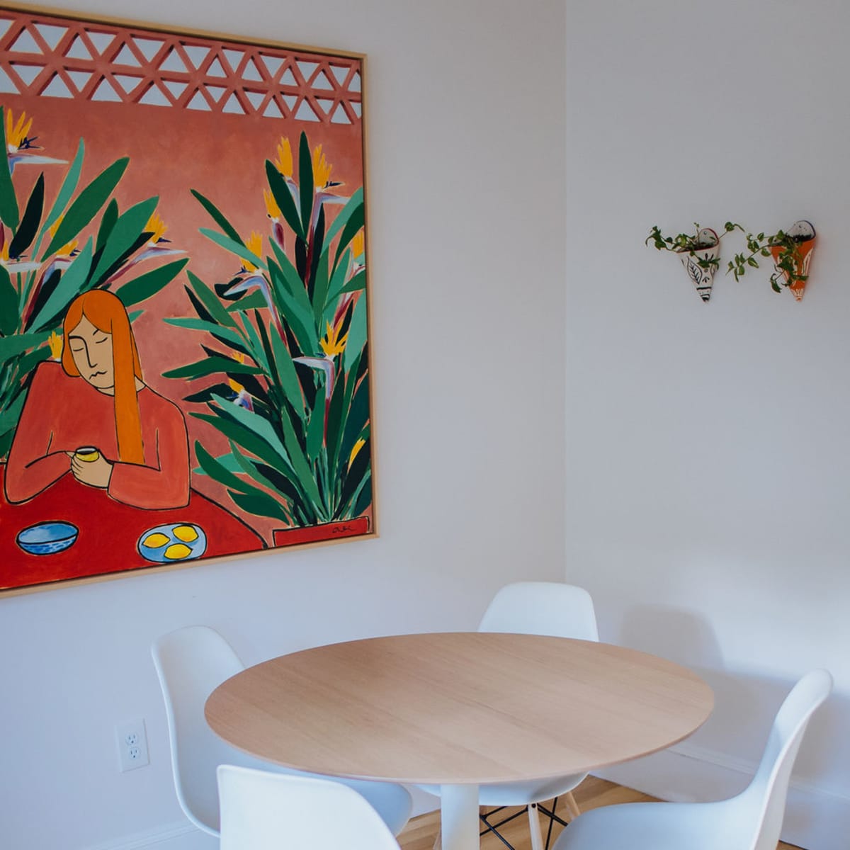 A round wooden dining table surrounded by four white chairs is positioned in a room featuring a colorful artwork on the wall.
