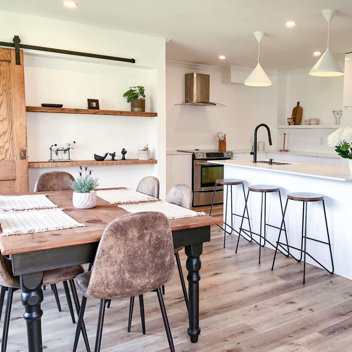 A modern kitchen and dining area features a white countertop island with bar stools, a wooden dining table, and shelves displaying decorative items.