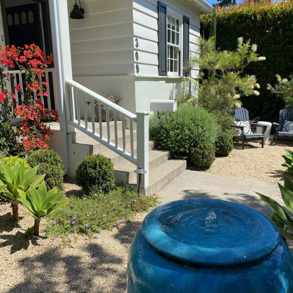 A blue ceramic fountain stands in a landscaped front yard with shrubs and flowering plants beside a white house.