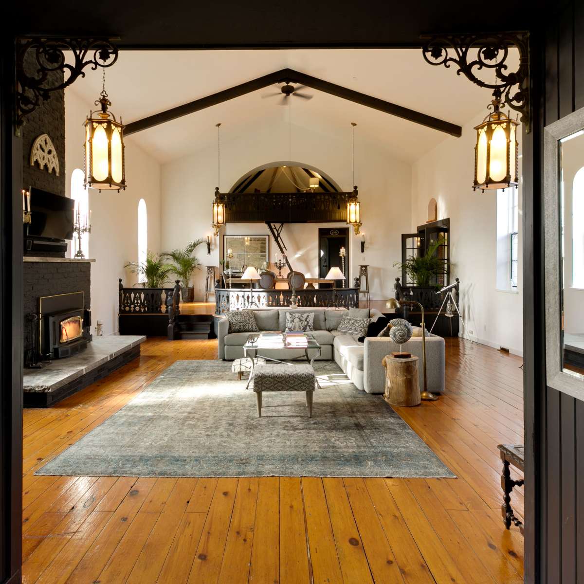 A large, open living room features a central seating area with a gray sofa and a coffee table, flanked by two decorative fireplaces and illuminated by hanging light fixtures.