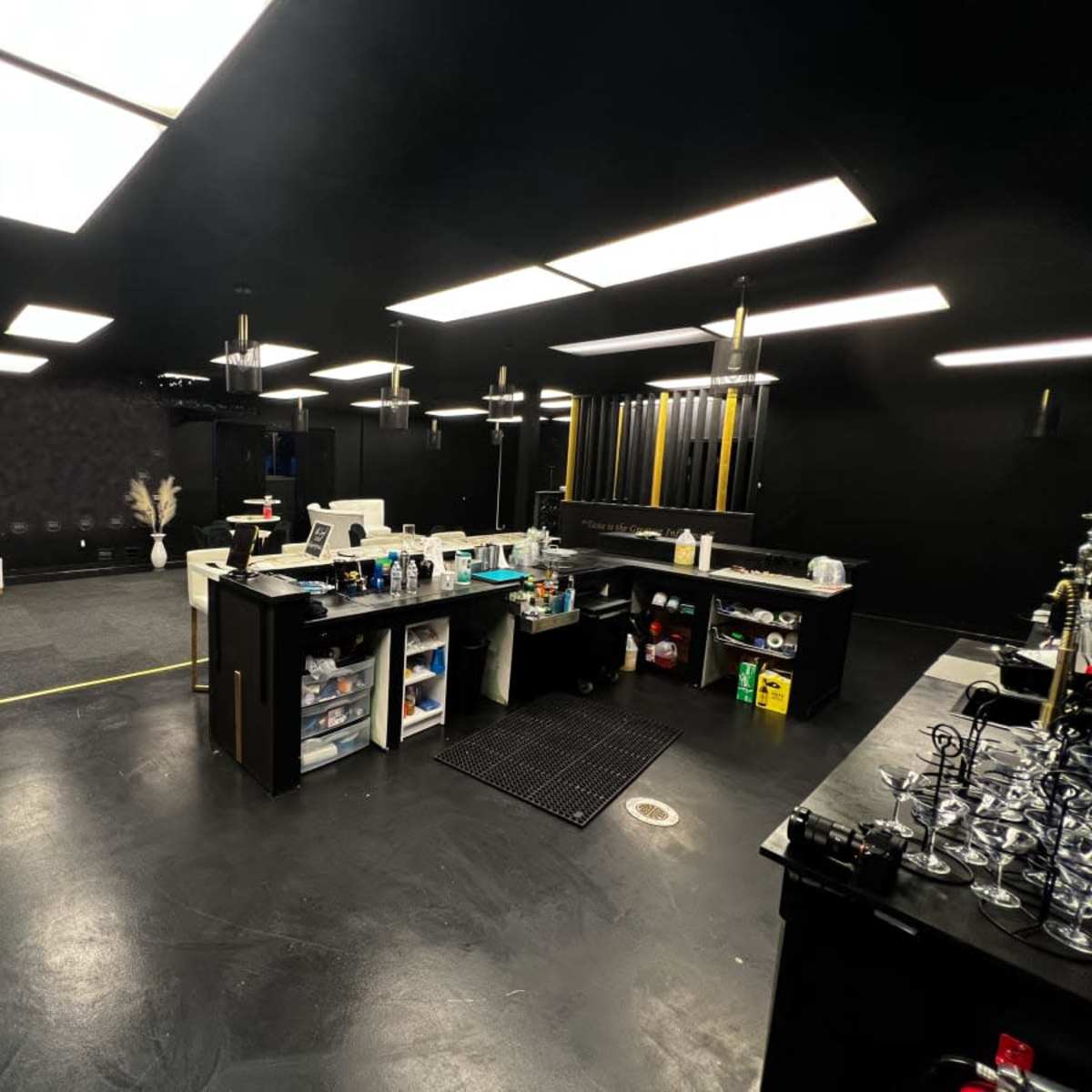 A modern, dark-themed workspace with black walls and flooring, featuring a bar-like counter with various items and tools organized on shelves and surfaces.